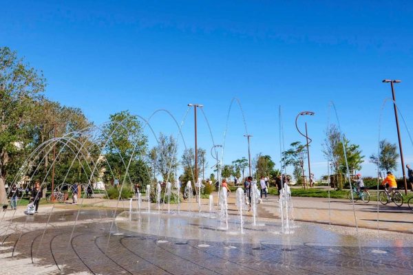 Fontane d'acqua in un parco cittadino con persone che passeggiano e alberi sullo sfondo.
