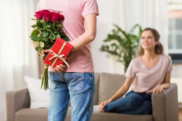 Una persona tiene un mazzo di rose e un regalo dietro la schiena, rivolto verso una donna sorridente seduta sul divano.